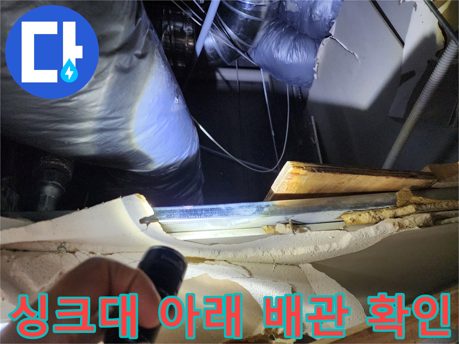 손전등으로 비춘 싱크대 하단부의 젖은 마감재와 배관 연결 부위