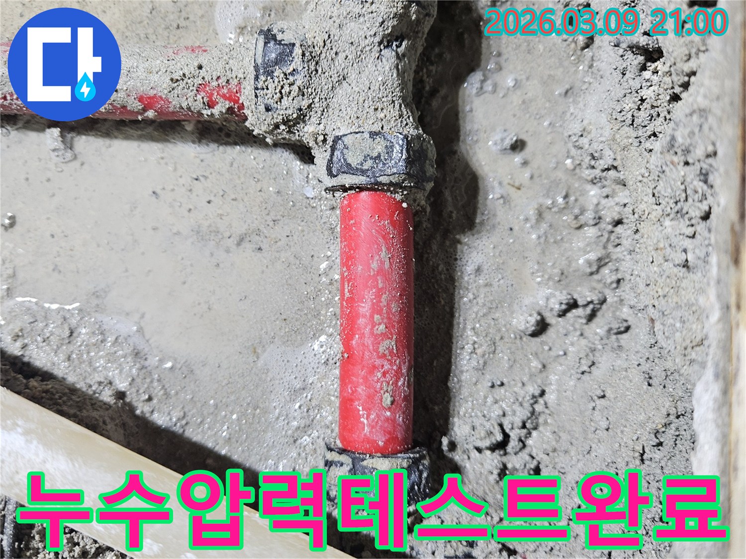 젖은 시멘트 바닥에-매립된 빨간색 배관의-누수 지점을-보수 후 압력 테스트를-진행하여-완벽하게 검증한-모습입니다