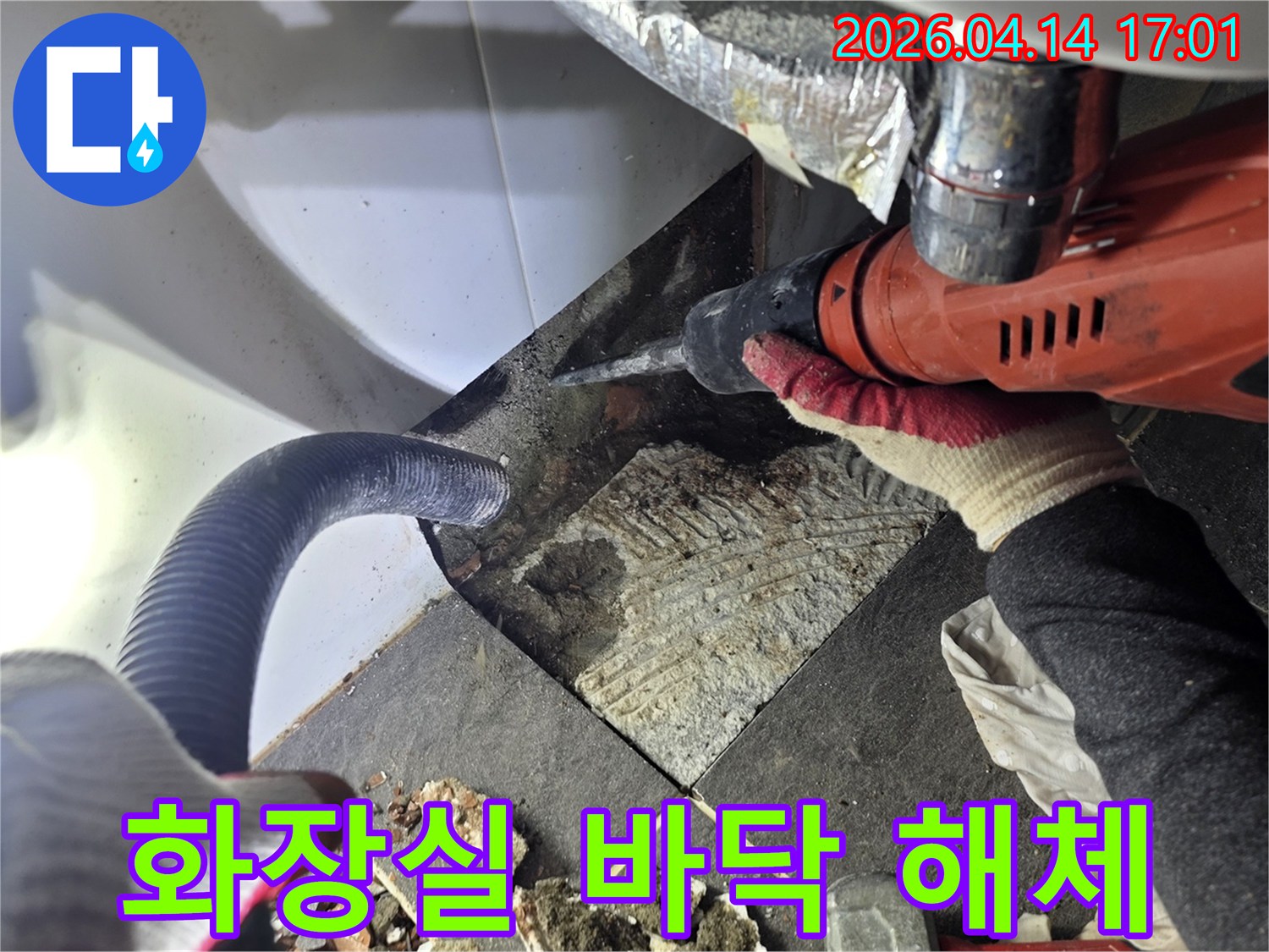 누수 지점의 바닥 타일을 해체하는 작업자가 드릴을 사용하며-파손된 타일 조각과 먼지 흡입 호스가 함께 보이는 작업 현장