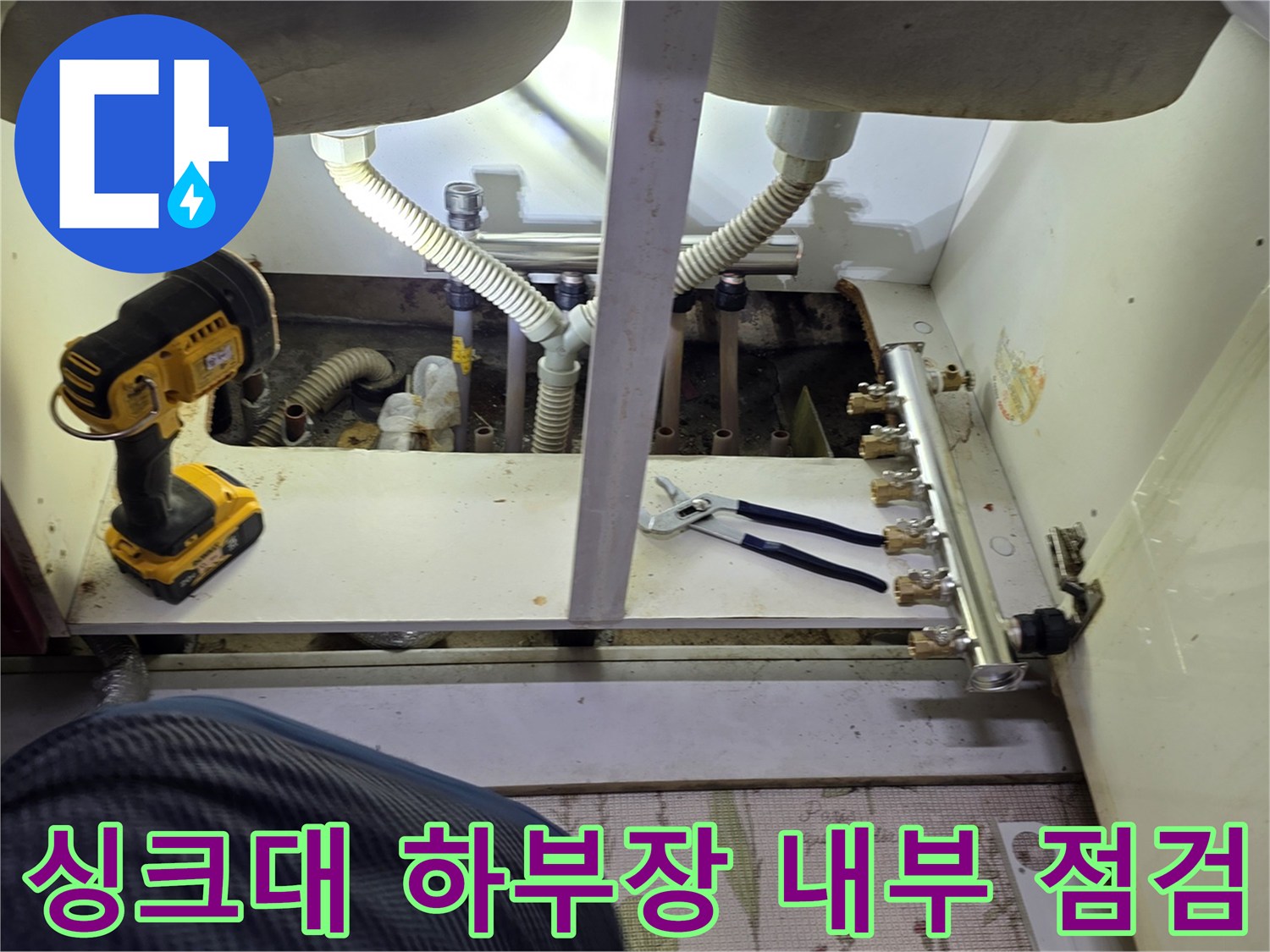 작업용 전동 드릴과 함께 싱크대 아래 분배기 배관을 점검하는 장면입니다
