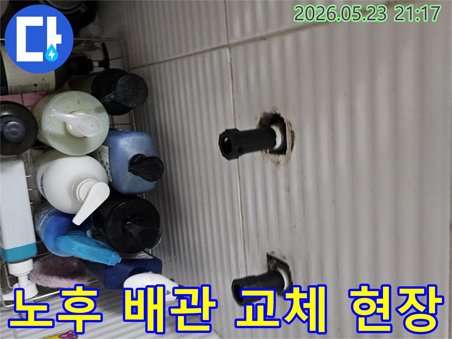 타일 벽면에서 노출된 오래된 수도 배관의 연결 부위를-누수로 인해 새 배관으로 교체하기 위해-점검하는 모습입니다.