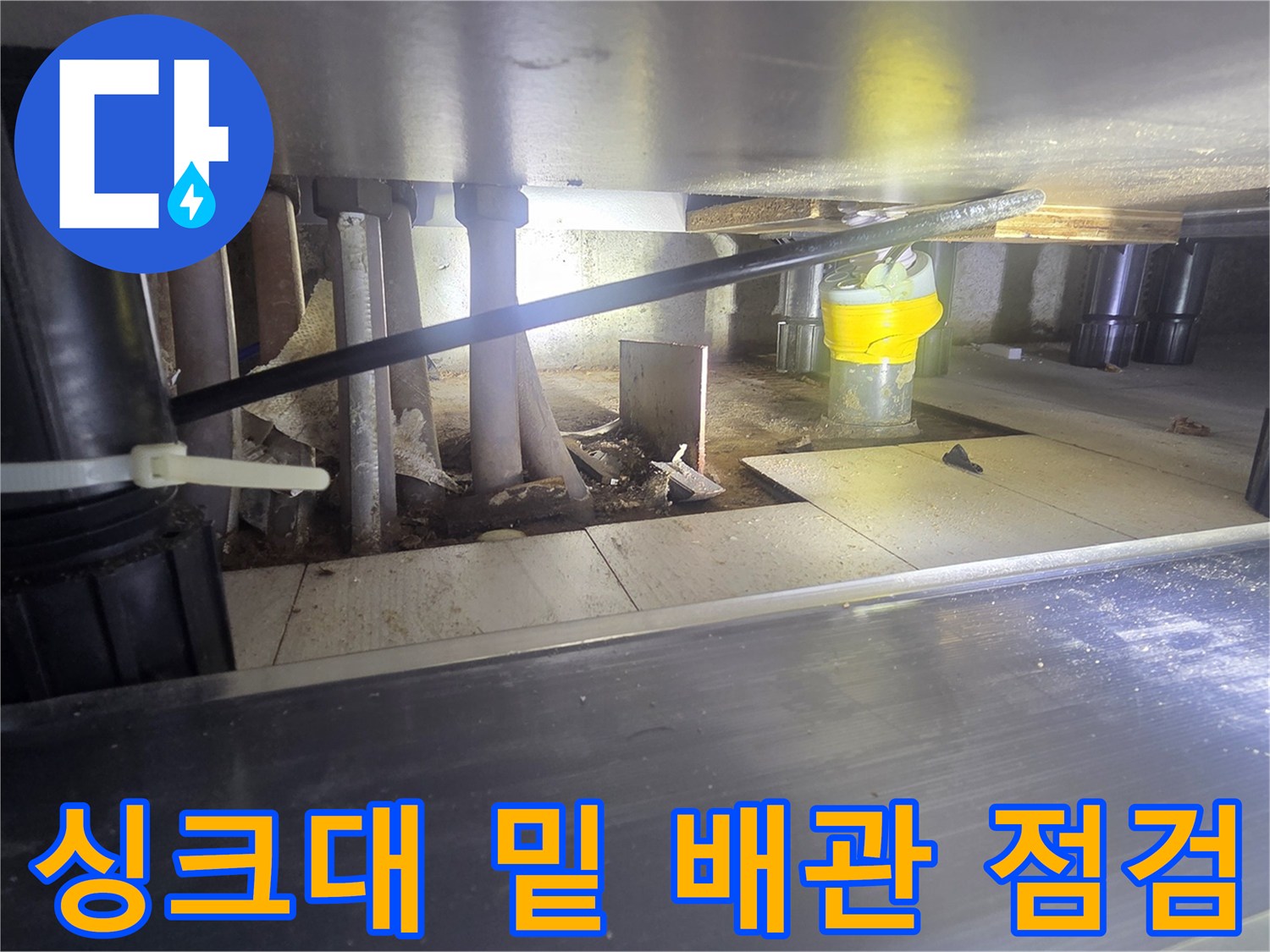 어두운 싱크대 하부 공간의 배관 연결 부위를 조명으로 비춘 모습
