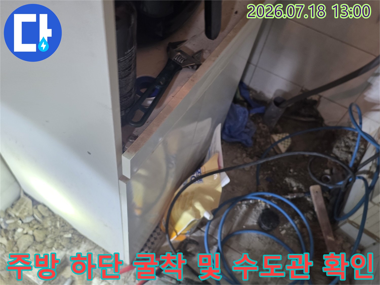 싱크대 아래쪽 타일이 제거된 자리에 파란색 배관과 빨간 집게 등 공구가 놓인 모습