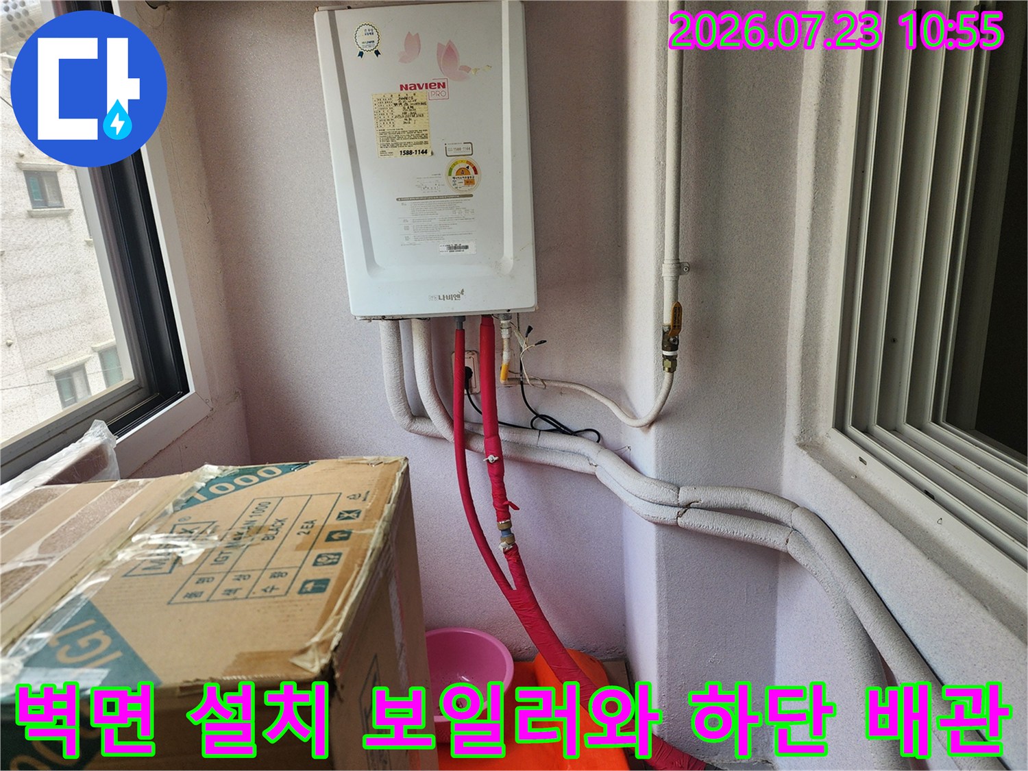 흰색 가스보일러 본체 아래로 붉은색 온수 배관이 수직으로 내려오며 바닥과 연결된 전경
