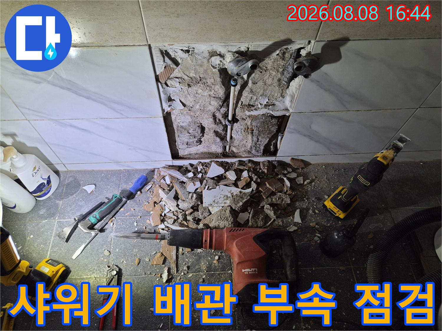 파쇄된 타일 사이로 드러난 샤워기 연결 배관과 작업용 드릴 장비입니다.