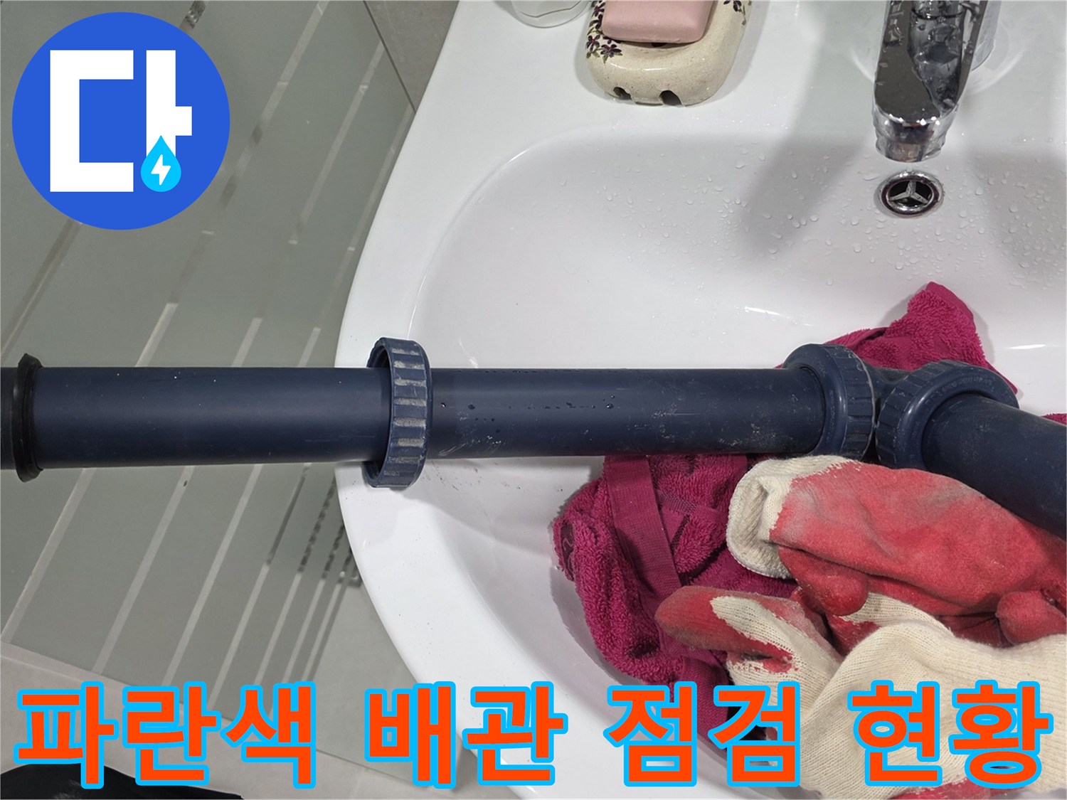세면대 아래 파란색 오배수관과 작업용 장갑이 놓여 있는 모습입니다.