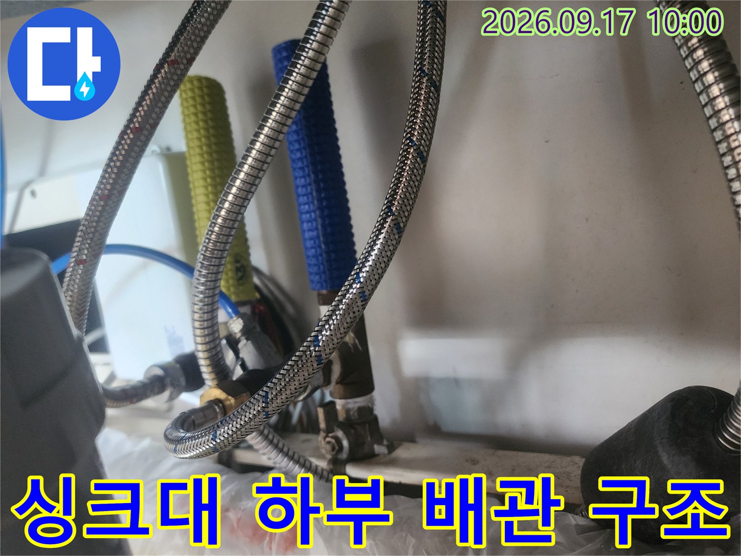 은색 메탈 호스와 파란색 보온재가 포함된 싱크대 밑부분의 복잡한 배관 연결 모습입니다.